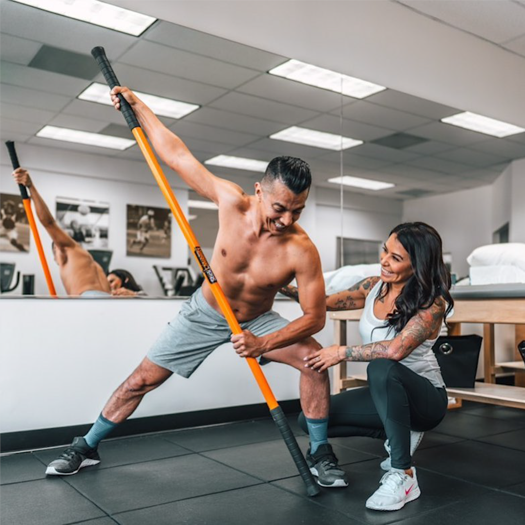 A female trainer providing hands-on guidance to a male client using a Stick Mobility training stick, focusing on proper technique for mobility and strength exercises.