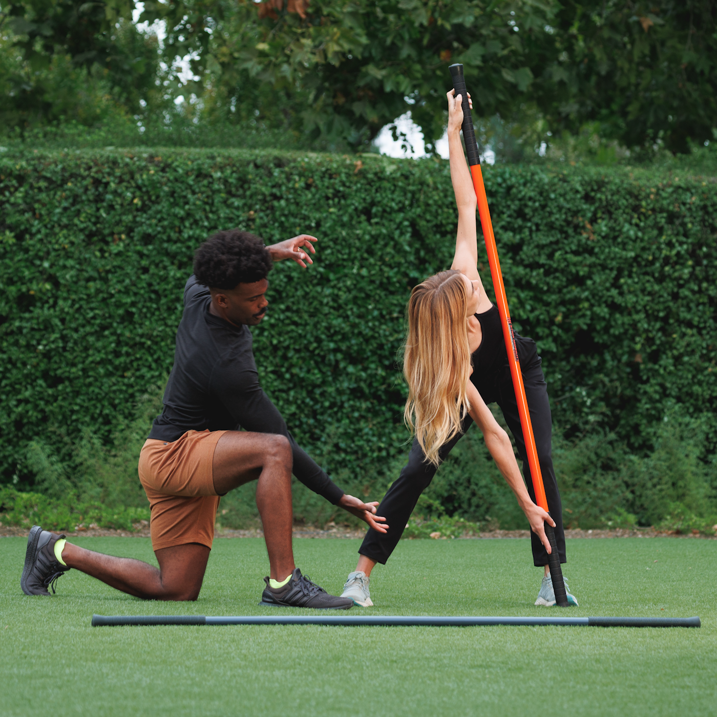 A male trainer outdoors coaching a female client on a back stretch using a Stick Mobility training stick, emphasizing proper form, mobility, and flexibility training.