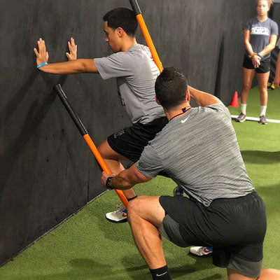 One man with his hands against the wall in a high knee position driving one knee towards to wall. Another man is using two training sticks to maintain posture and technique for the guy performing the movement.
