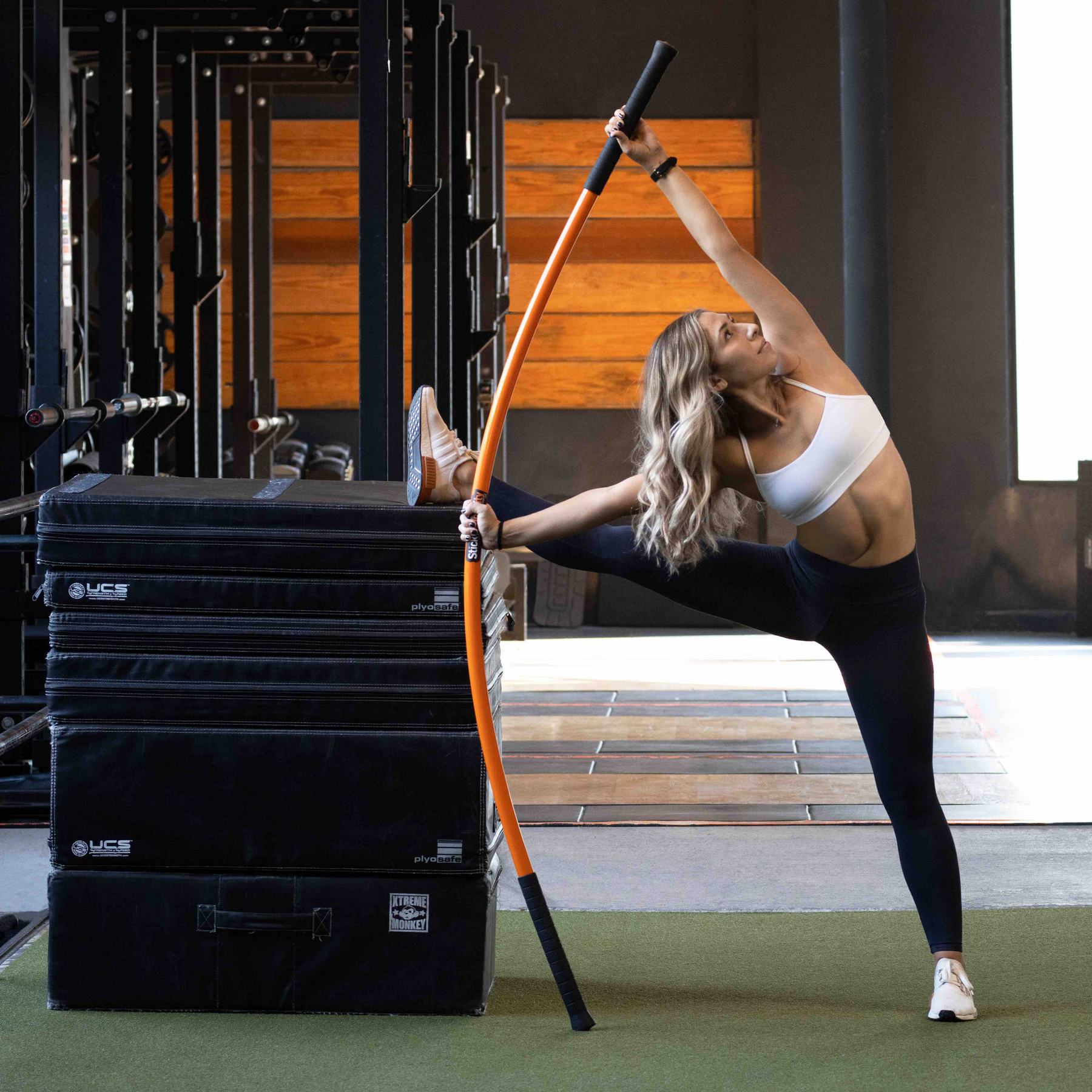 Woman with one leg elevated on boxes, while performing the Stick Mobility bow and arrow.