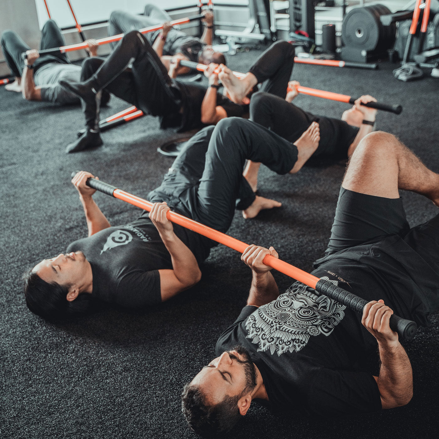 Two men laying on the ground in a glute bridge position while each holding an end of the Training Stick above their chest.
