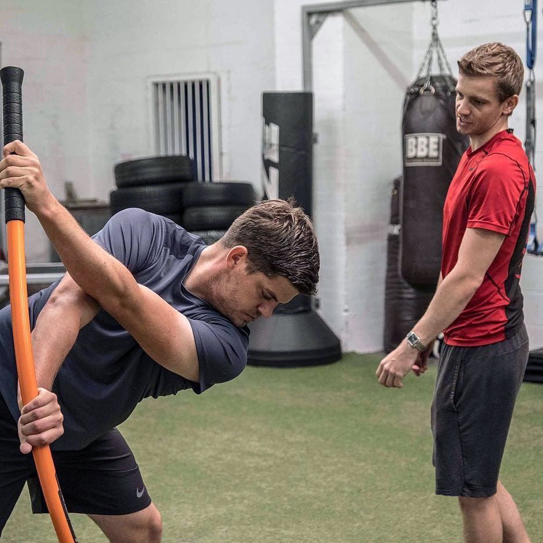 A man stretching with the Training Stick while another man provides feedback.
