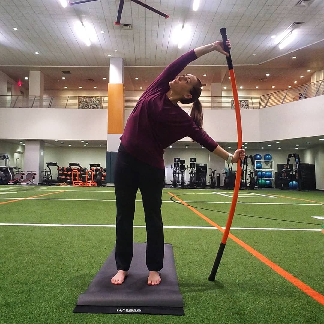 A woman stretching with a Training Stick bending to the side while standing on a mat.