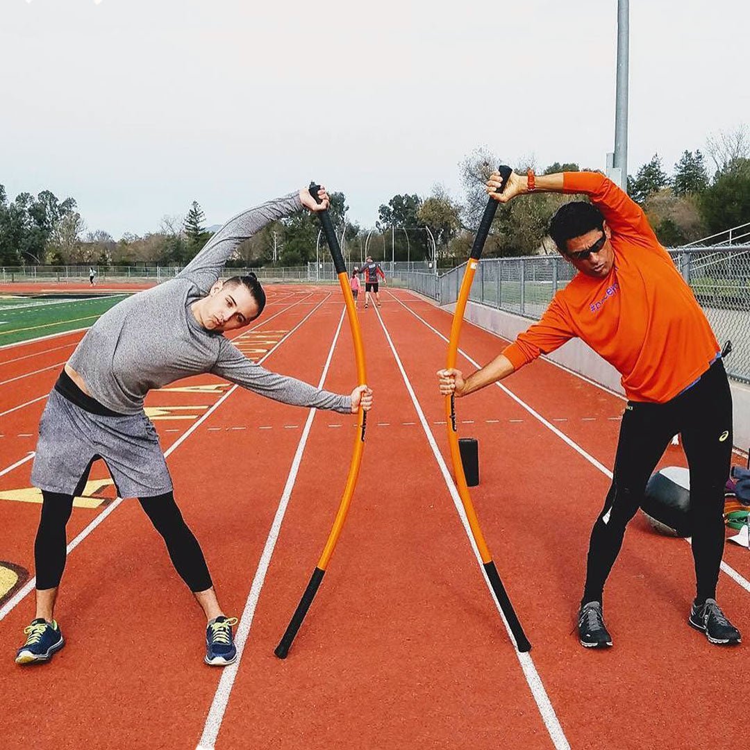 Two men stretching with Training Sticks,  bending them to the side, while on a track.