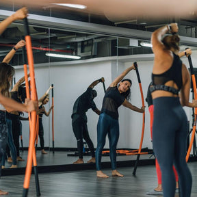A group of women from MEFITPRO practicing the bow & arrow stretch with Stick Mobility training sticks in a gym, highlighting mobility, flexibility, and group fitness dynamics.