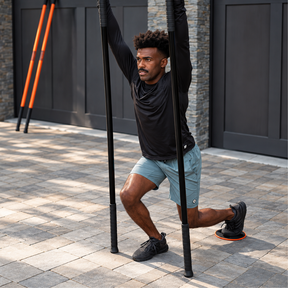 A man in a black long-sleeve shirt and light blue shorts performs a lunge outdoors while using Stick Mobility Training Sticks. 