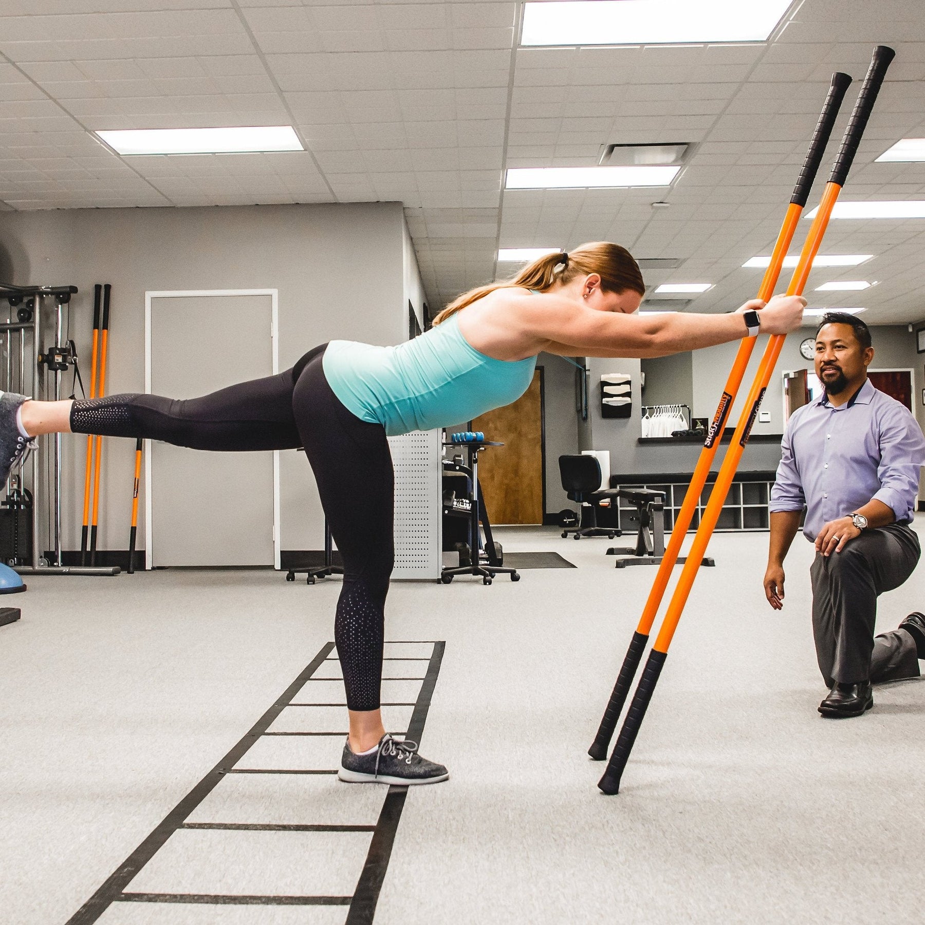 Woman performing a single leg hinge while holding on to two Training Sticks while a man watches her form. 