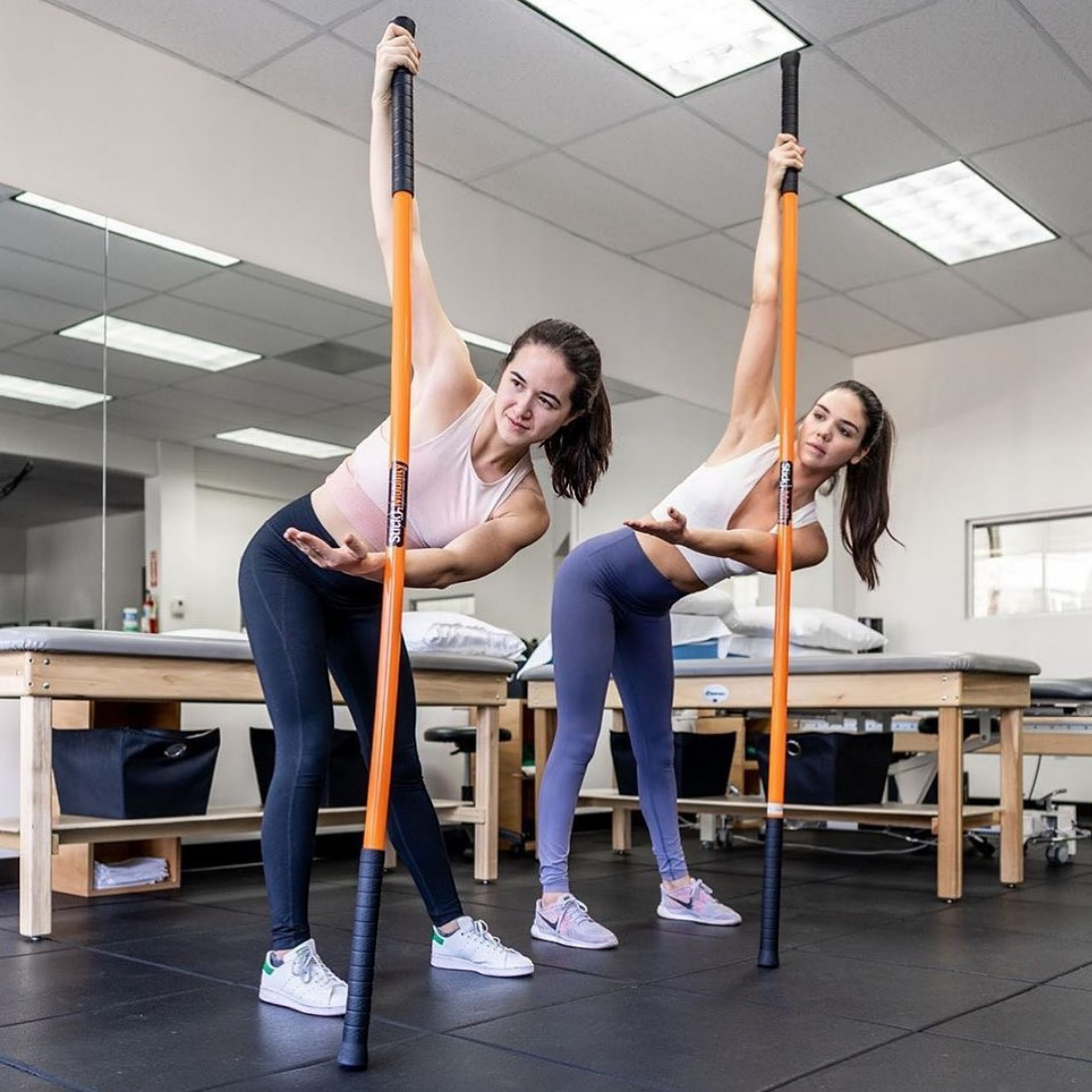 Two women performing a hip hinge and twist with the Training Stick.