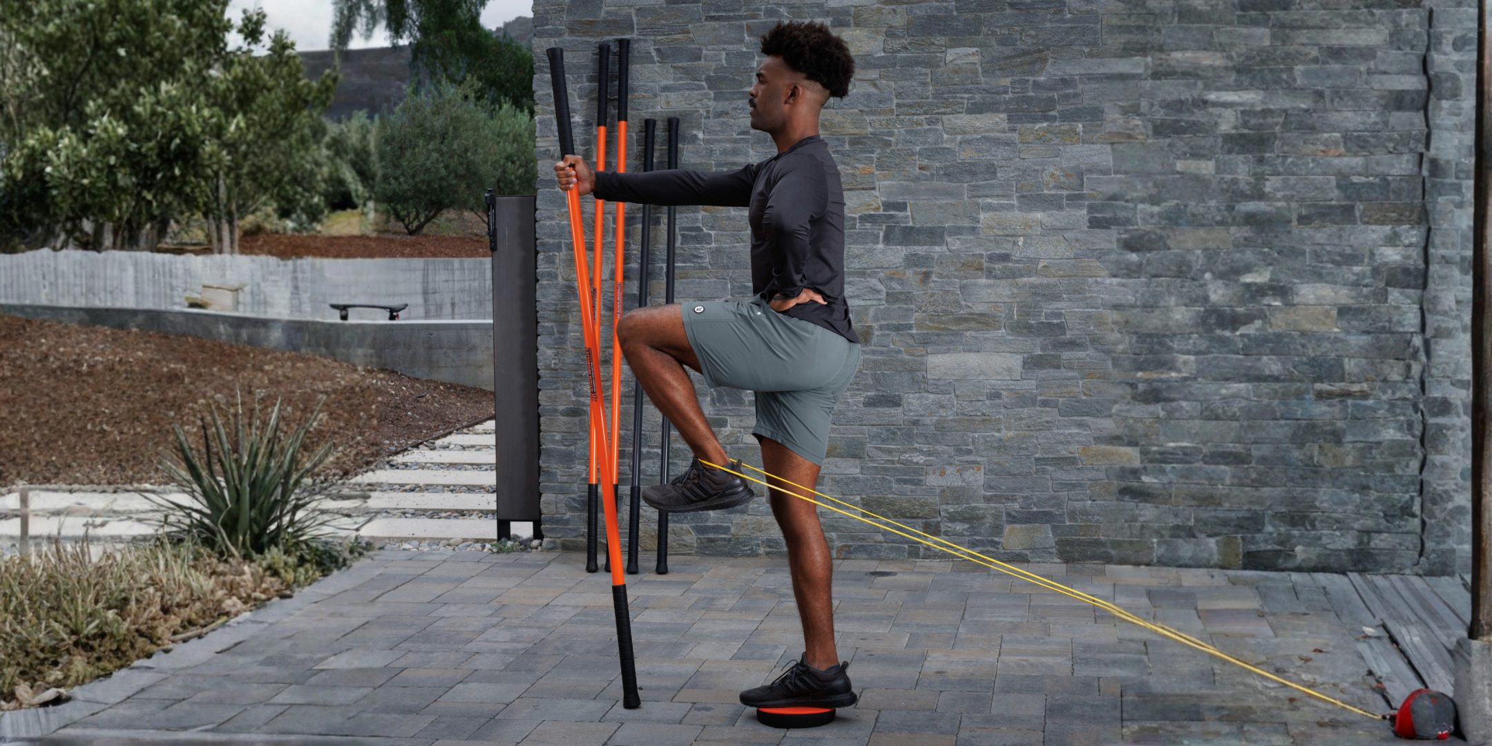 A man performing an exercise outdoors using a resistance band and a vertical stability rod on a paved patio in front of a gray stone wall.